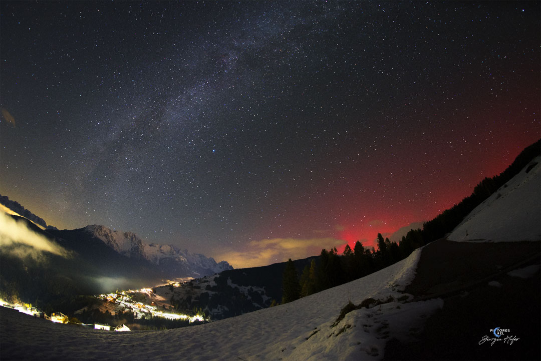 The night sky over a valley is shown complete with the central band of the Milky Way Galaxy crossing up from the lower left. On the right the sky just over the hill glows an unusual red: aurora. Please see the explanation for more detailed information.