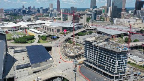 An image of a large construction project in the middle of a hole in the ground next to a large futuristic looking stadium in Atlanta near many tall buildings.