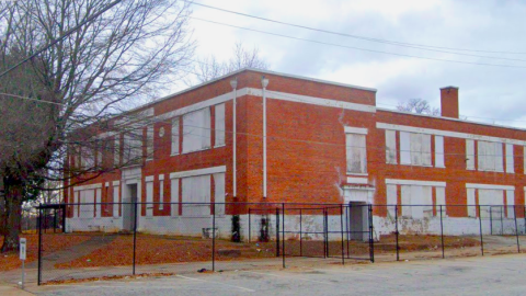 An image showing an old brick school in Atlanta with boarded-up windows and a wide parking lot next door, under gray-blue skies.