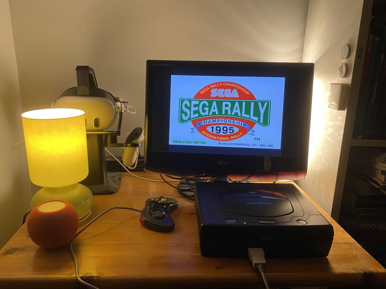 A small table with a 20-ish inch TV and a Sega Saturn running Sega Rally Championship 1995. A Quest 3 can be seen in the background on its charging stand.