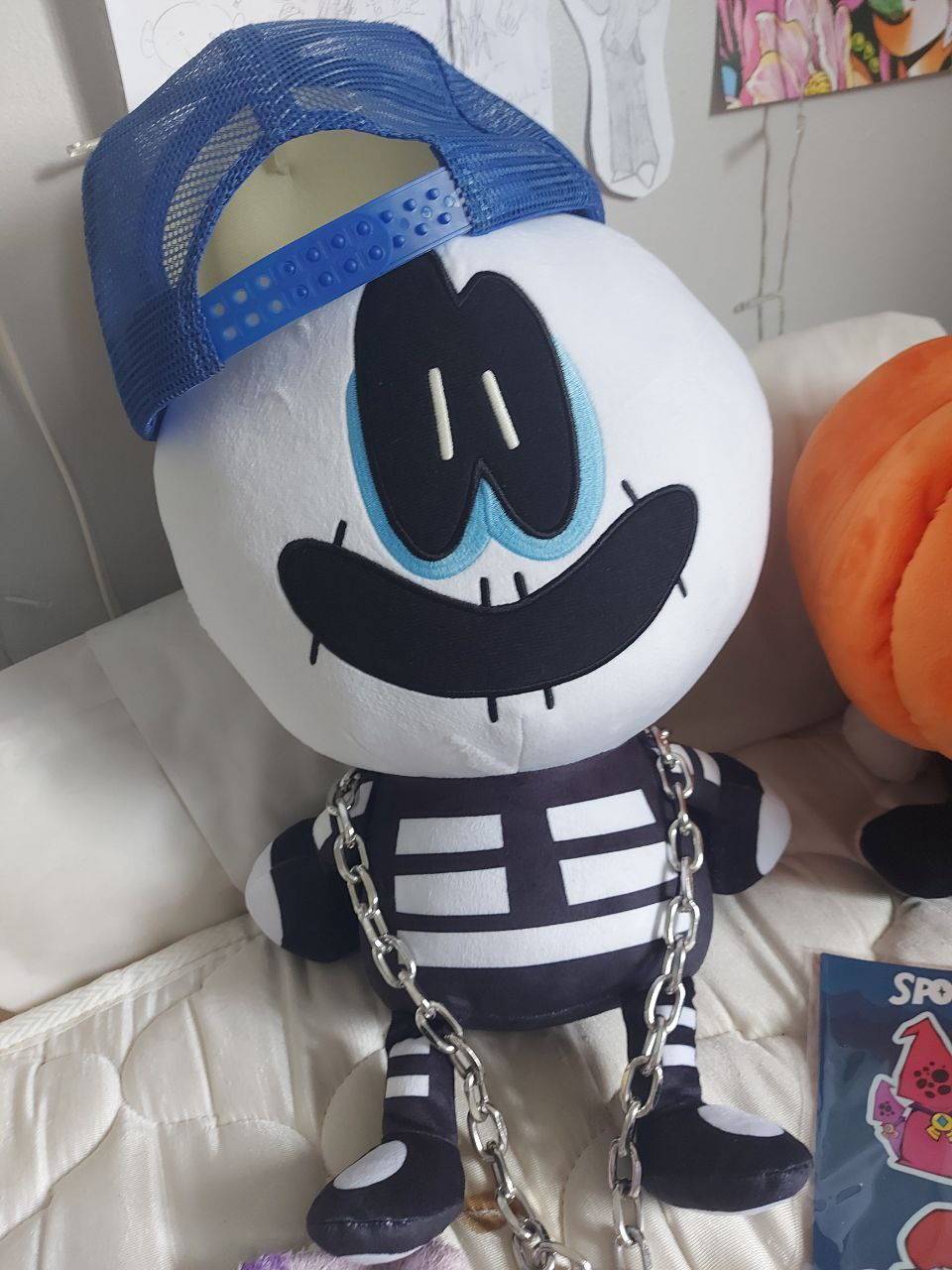 A plush doll of Skid from the YouTube series Spooky Month, sitting on a bed with no sheets on it. My son is an absolute fucking monster and refuses to change his sheets. What the fuck, bro? Just put the sheets on. It's not hard. Anyway Skid is wearing a metal chain and a backwards ball cap that Dipper from Gravity Falls wears. Pump is off frame I guess. CHANGE YOUR FUCKING SHEETS DUDE HOLY SHIT.