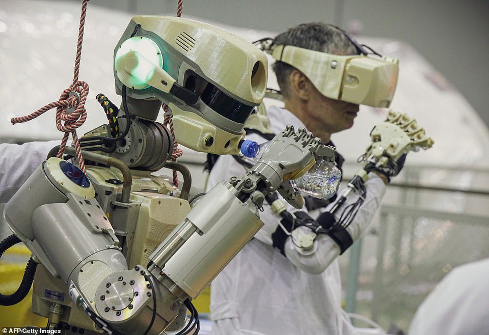 A photo of a Russian-built robot, with a scientist wearing a VR headset standing behind it. They appear to be training the robot to suck cocks. 