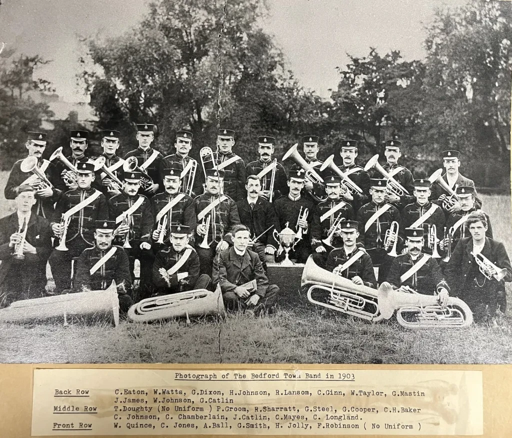 A black and white photograph of Bedford Town Band in 1903. A group of men holding various brass instruments in uniform with hats, in a field with trees behind them. The bottom of the photograph lists the names of everyone in the back row, middle row, and front row